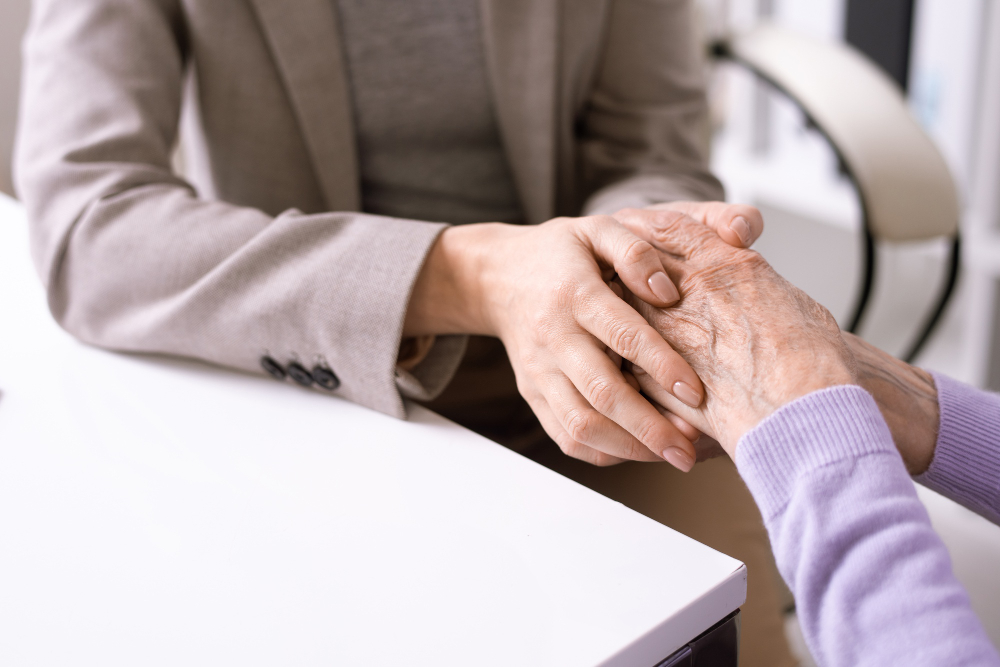 Gros plan sur deux paires de mains posées sur une table blanche, une personne en veste beige tenant avec bienveillance les mains ridées d'une personne âgée en pull mauve