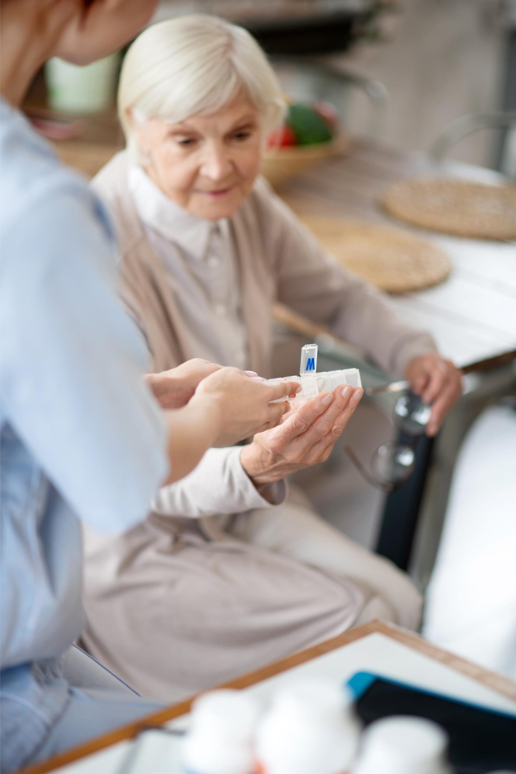 Une auxiliaire de vie tend un pilulier ouvert à une femme âgée assise à une table de cuisine, un verre d'eau à la main